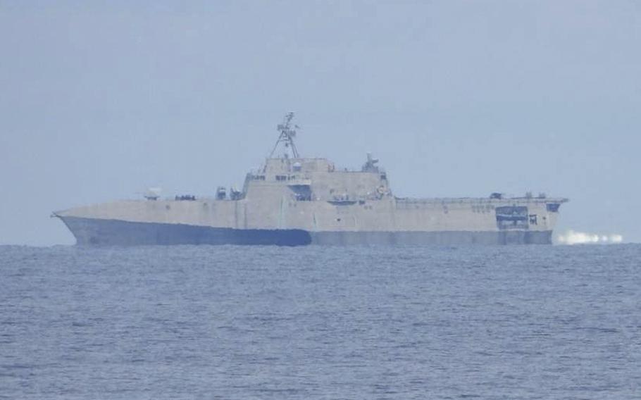 A naval warship, seen from a distance, sails in open water with the hazy horizon in the background.