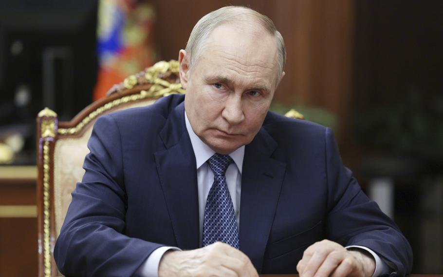 Vladimir Putin, wearing a dark suit, sits at a table with his hands in front of him holding a pen.