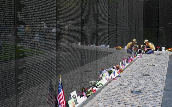 A woman in a yellow shirt and hat kneels down to place flags at the base of a black marble wall inscribed with names.