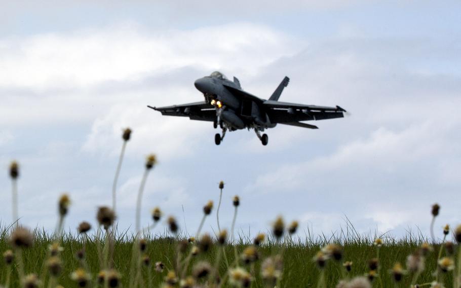 A military fighter jet descends above a grassy field to land with cloudy blue sky behind it.