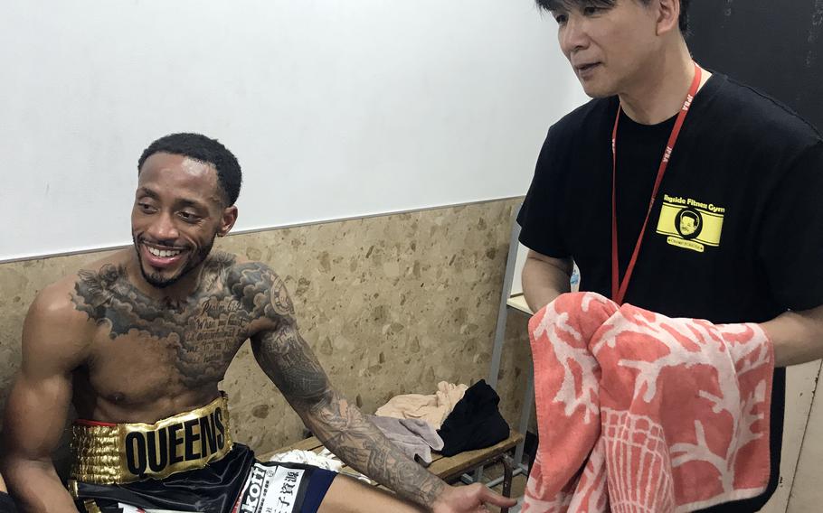 A boxer in black shorts smiles while sitting on a bench in a locker room as a trainer in a black shirt stands next to him.
