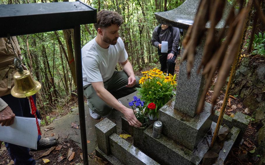 A man in a white shirt kneels to place flowers on a marble shrine in a forested area.