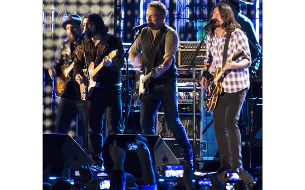 Several people perform on stage, four in the front with guitars and one in the back who is mostly obscured.