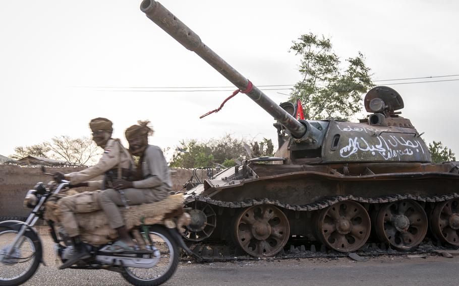 RSF soldiers ride a motorcycle on a main street in El Geneina past a destroyed tank that belonged to the Sudanese military.