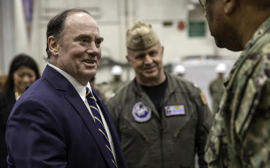 A man in a suit speaks with two military members in camouflage uniforms in a hangar bay.
