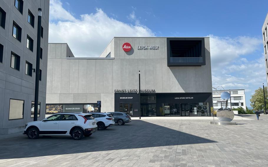 A modern concrete building houses the Ernst Leitz Museum with prominent red Leica logo and “LEICA-WELT” signage on the facade. The contemporary architectural structure features clean geometric lines, large windows, and houses both a museum shop and Leica Store Wetzlar. Several cars are parked in front of the building on a paved plaza area.