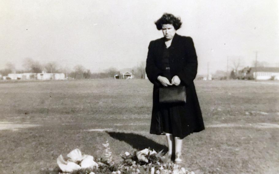 In a black and white photo, a woman dressed in dark clothing stands solemnly next to a grave covered in flowers.