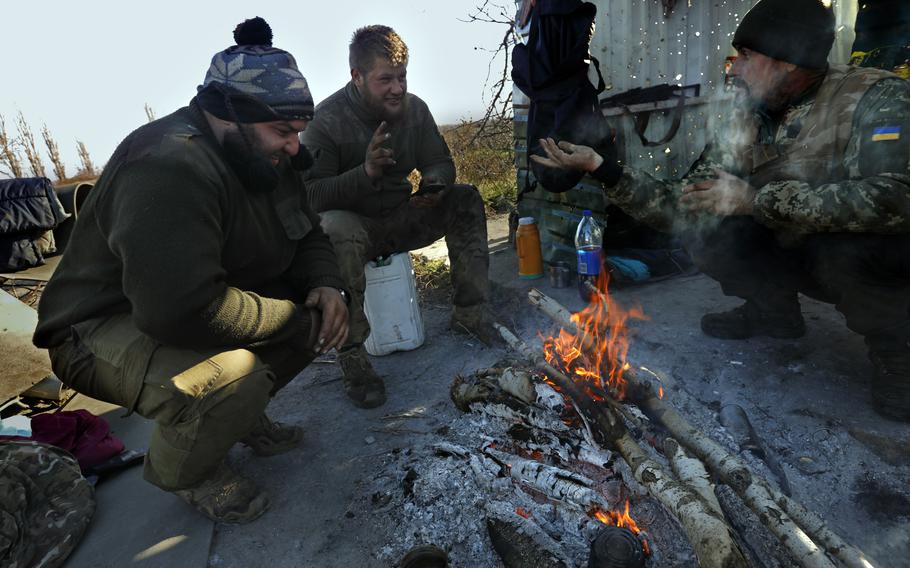 Members of a Ukrainian army tank platoon get warm by a fire beside a bus stop riddled with bullet holes.