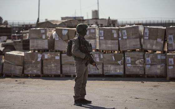 A soldier in camouflage combat gear and holding a machine gun stands on a concrete surface next to a large stack of cardboard boxes.