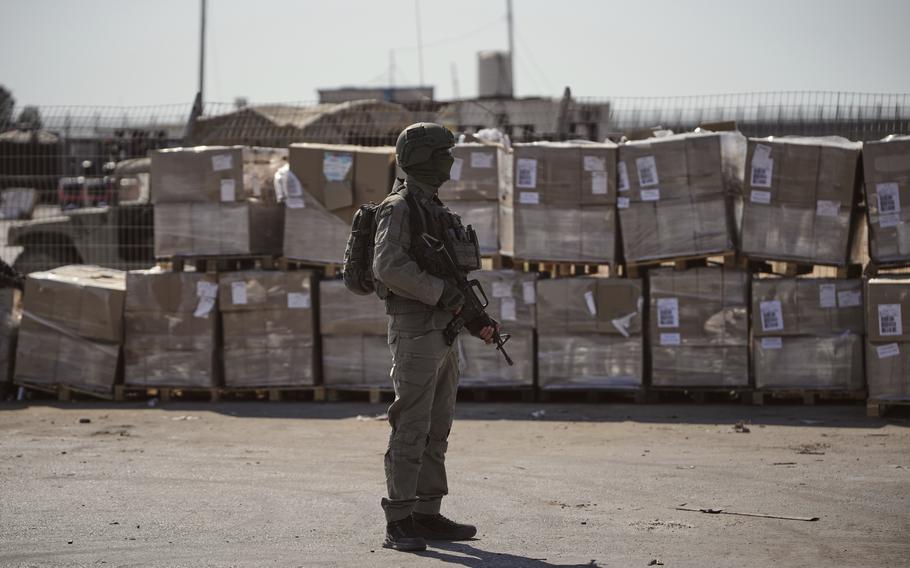 A soldier in camouflage combat gear and holding a machine gun stands on a concrete surface next to a large stack of cardboard boxes.