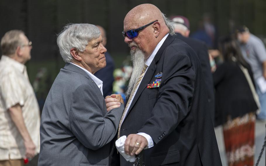 Two men in suits shake hands as others mingle behind them along a black memorial wall.