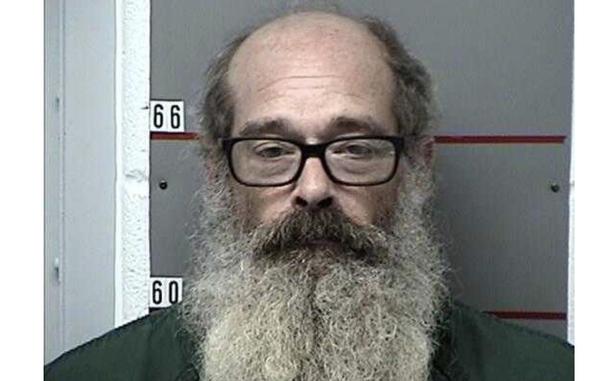 A police booking photo of a man balding on the top of his head with black-rimmed glasses and a long gray beard.