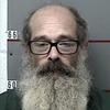 A police booking photo of a man balding on the top of his head with black-rimmed glasses and a long gray beard.