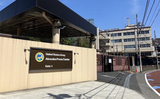 An brown entrance sign on a wall reads “United State Army Akasaka Press Center” next to a gate outside a military building.