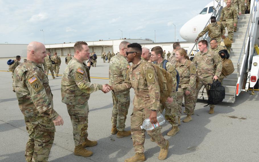 Soldiers shake hands with leaders standing to receive them as they walk off the steps of a plane.