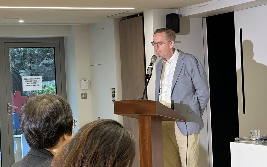 A man in glasses and a light blue blazer speaks at a wooden podium with a microphone in a conference room with audience members visible in the foreground and a window showing greenery in the background.