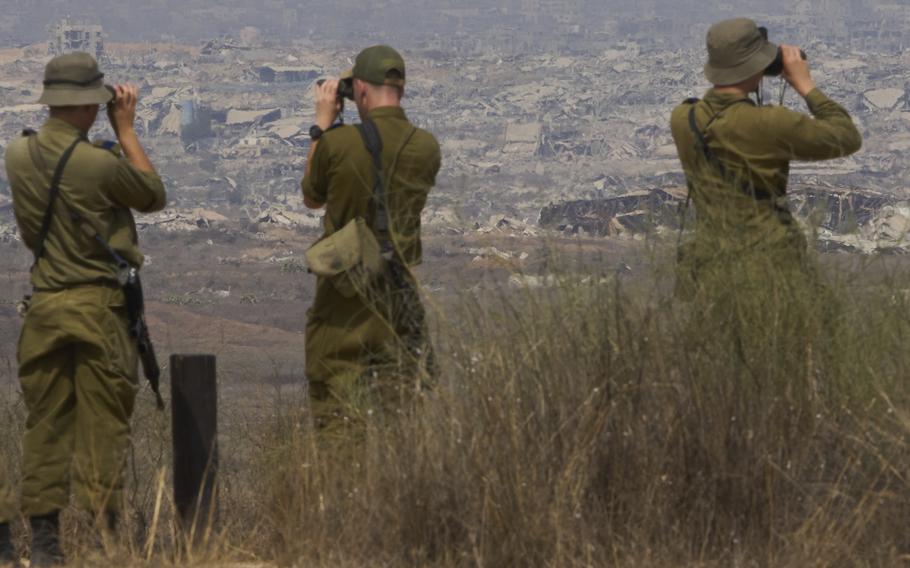 Three soldiers in green uniforms, seen from behind, use binoculars to observe a cityscape of damaged buildings in the distance.