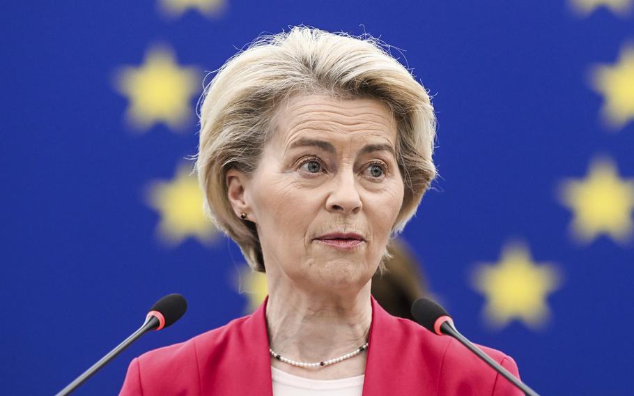 A woman with short blonde hair wearing a red suit speaks into two mircrophones while standing in front of a blue flag with a circle of gold stars on it.