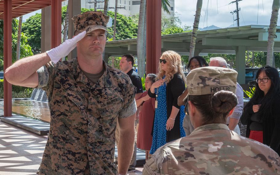 A U.S. Marine in desert camouflage uniform and cover salutes. The scene takes place under a covered outdoor pavilion with palm trees visible in the background. Other service members in various uniforms and civilians, including a woman in a blue polka dot dress, are gathered in the background participating in the solemn ceremony.