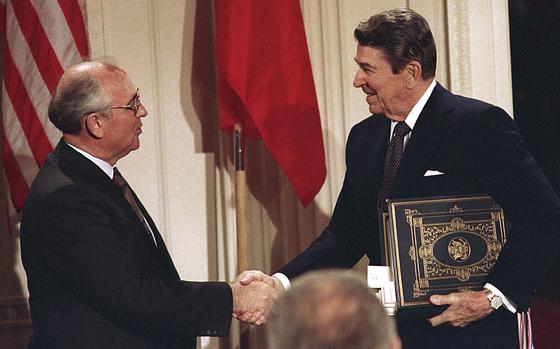 Ronald Reagan holds a folder tucked under his arm and shakes hands with Mikhail Gorbachev in front of U.S. and Russian flags.