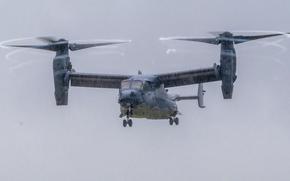 A V-22 Osprey tiltrotor aircraft flies through overcast skies with its twin rotors in horizontal position for forward flight. 