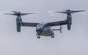 A V-22 Osprey tiltrotor aircraft flies through overcast skies with its twin rotors in horizontal position for forward flight. 