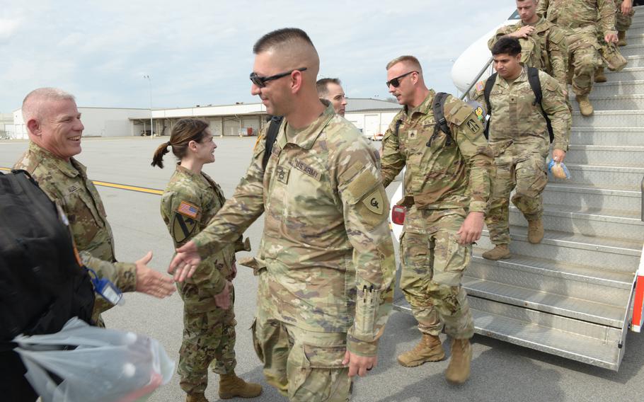 A line of soldiers walking off the steps of a plane and shaking hands with leaders standing to receive them.