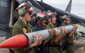 Four men in military flight suits, helmets and ear protection carry a large missile to a fighter jet on a runway.
