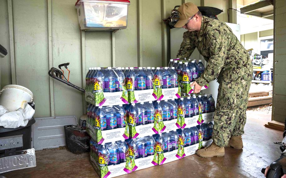 A military service member in camouflage uniform stacks cases of bottled water in front of a green-colored wall.