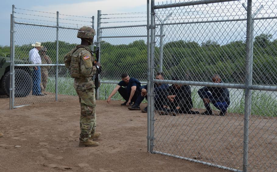 A Texas National Guard soldier watches over four migrants who entered a privately owned pecan orchard in Eagle Pass, Texas, after illegally crossing the Rio Grande from Mexico on May 23, 2022. The men were later processed by U.S. Border Patrol agents.