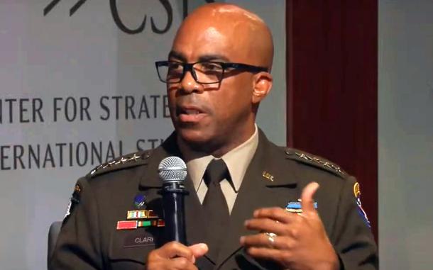 An Army officer in green dress uniform and black-rimmed glasses speaks into a microphone held in one hand while gesturing with the other hand.
