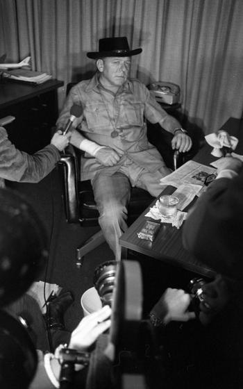A man sits in a chair while a microphone is held to him by another person next to him.