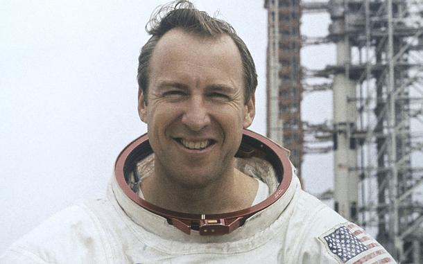 A 1970s-era image of a man in an astronaut’s space suit, seen from the neck up, with a metal launch tower in the background over his left shoulder.