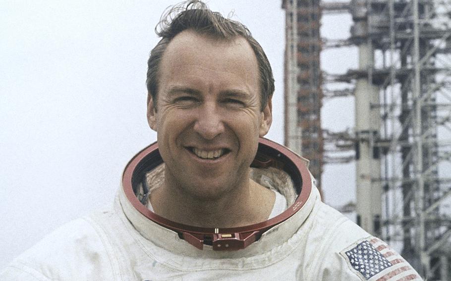 A 1970s-era image of a man in an astronaut’s space suit, seen from the neck up, with a metal launch tower in the background over his left shoulder.