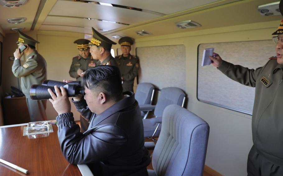 Kim Jong Un sits down and looks through binoculars as North Korean military members look on. 