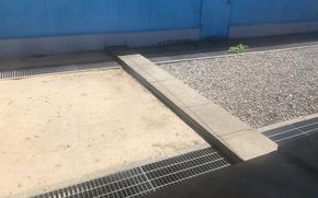 A narrow slab of concrete, seen from above, marking the border between South and North Korea sits across a walkway with gravel on one side, dirt on the other and a blue wall behind it.