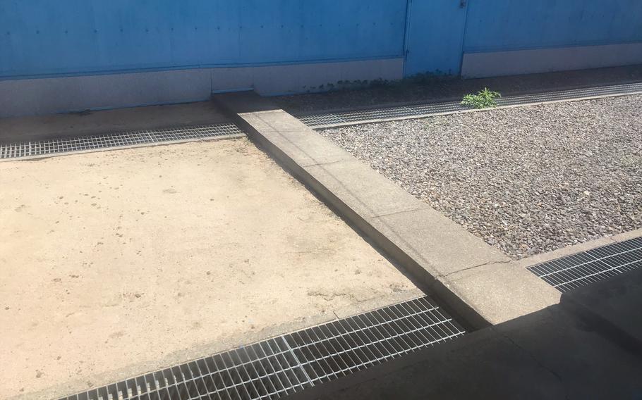A narrow slab of concrete, seen from above, marking the border between South and North Korea sits across a walkway with gravel on one side, dirt on the other and a blue wall behind it.
