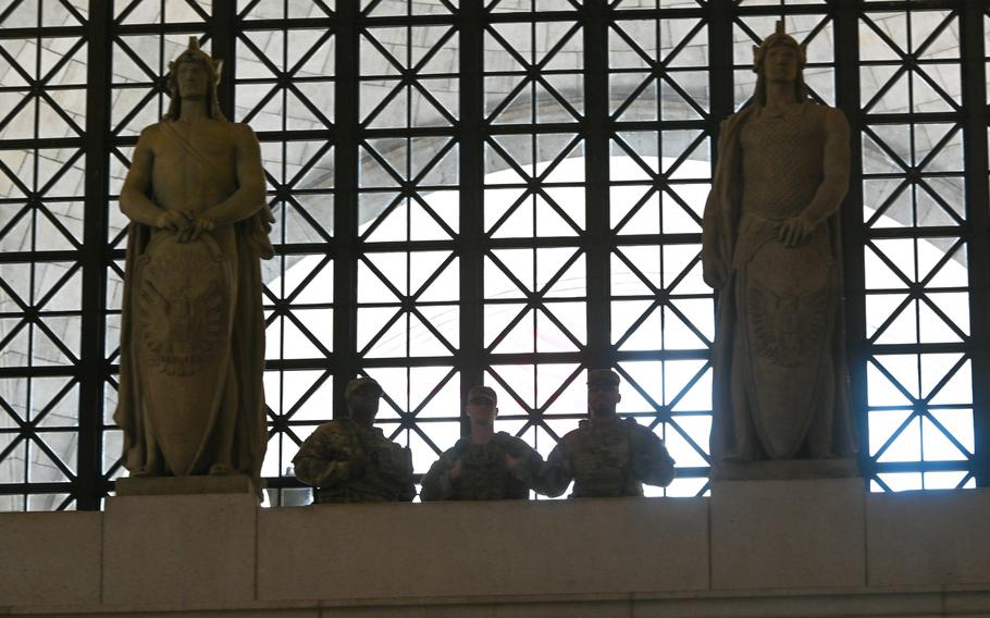 Three soldiers seen from the waist-up between two large statues; they are nearly silhouettes because of the bright light behind them.