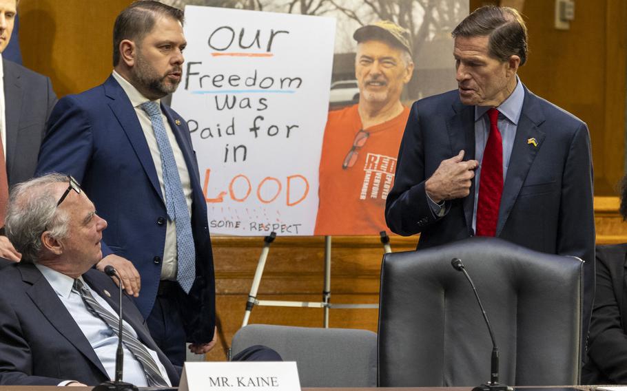 Three senators — one sitting, two standing — chatting with a poster in the background of a man holding a sign that reads, “Our freedom was paid for in blood.”