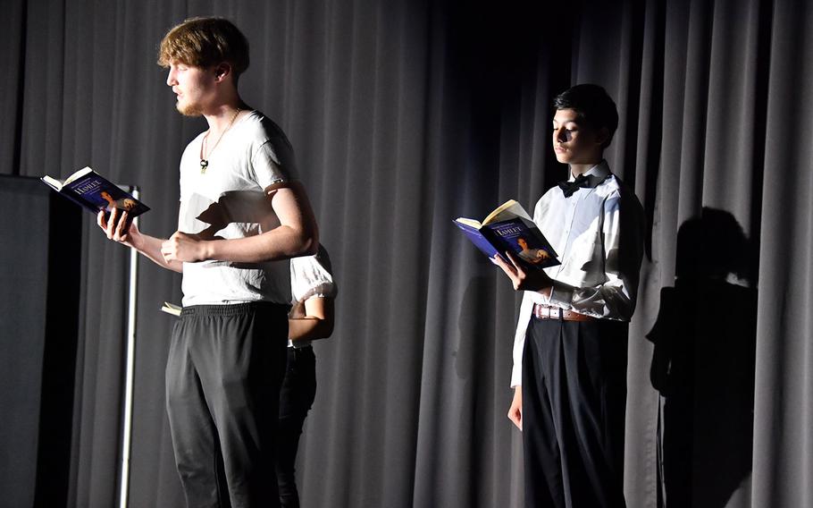Three teenagers, each wearing black pants and a white top, stand on stage reading from copies of Hamlet.