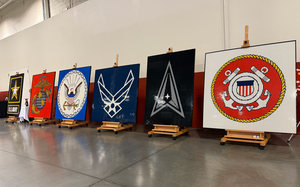 The logos of the six U.S. military services made out of logos are displayed on easels in front of a white and red wall.