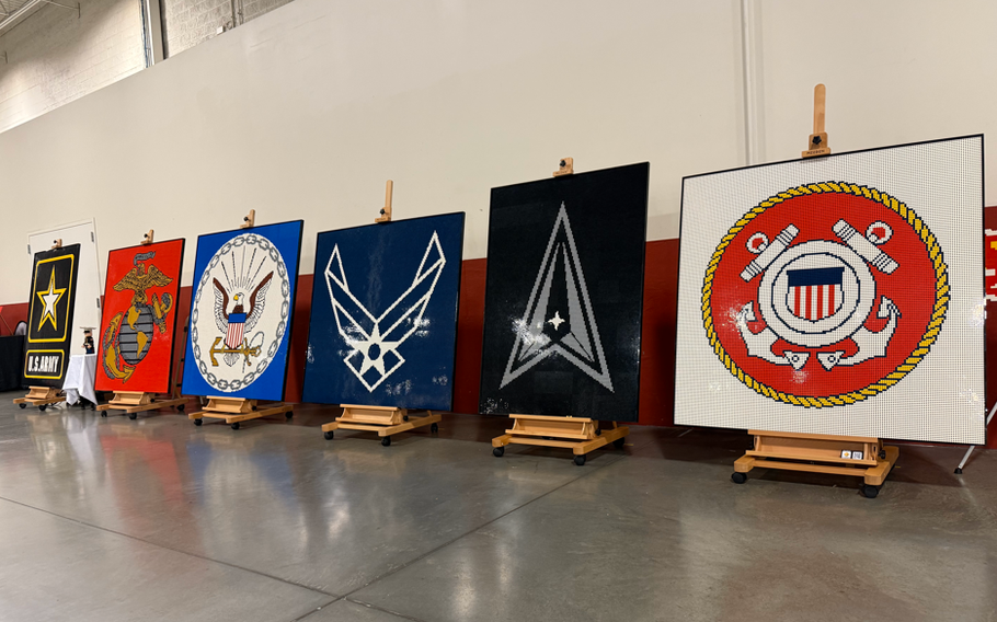 The logos of the six U.S. military services made out of logos are displayed on easels in front of a white and red wall.