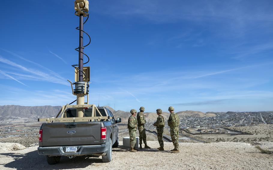 Army soldiers look at the border wall next to a surveillance vehicle.
