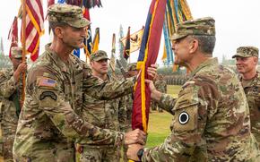 Two men in Army uniforms hold a small flag, as others in uniform look on near them.