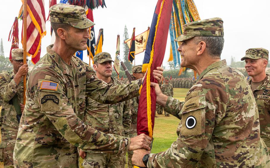 Two men in Army uniforms hold a small flag, as others in uniform look on near them.