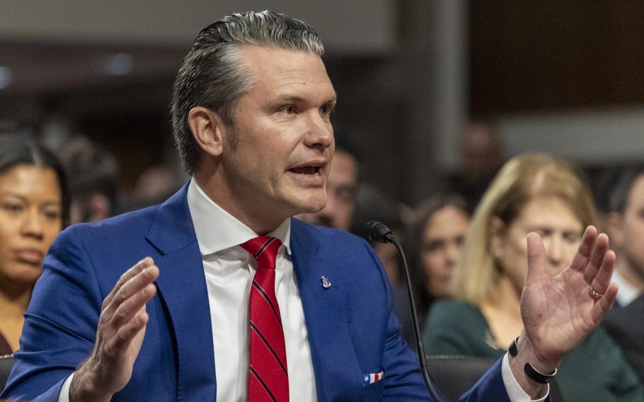 Pete Hegseth raises his hands while speaking into a microphone at his seat.