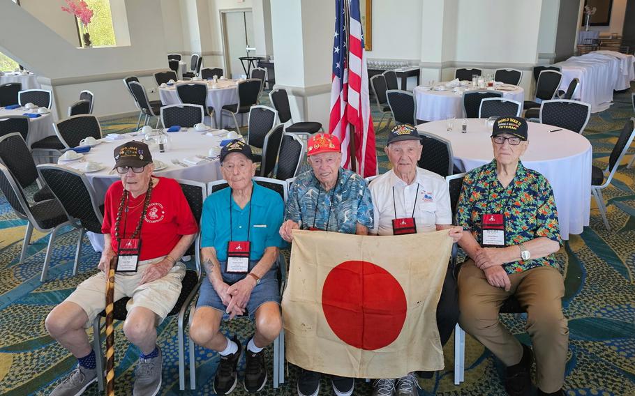 Five WWII veterans wearing baseball caps sit in chairs in a banquet hall. 