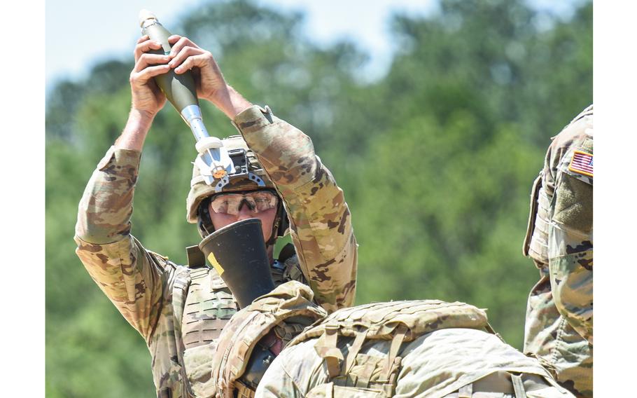 A soldier carries a mortar round with both hands, holding it above a launcher.