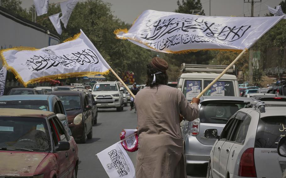 A man in the middle of a street holding several flags as cars pass by.