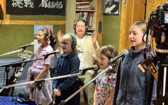 Children sing in front of microphones while their teacher directs them.